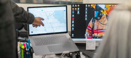 Two people at dual monitors, one points at a map.