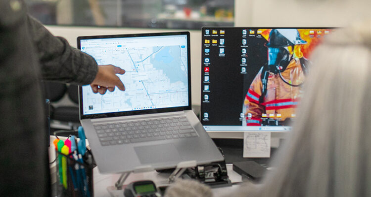 Two people at dual monitors, one points at a map.