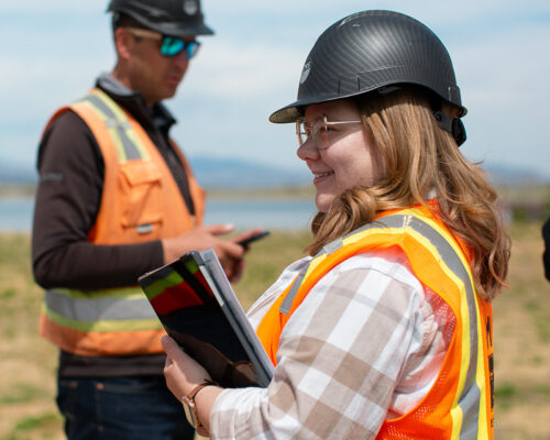 Two people in safety gear outdoors; one smiles with tablet, one on phone.