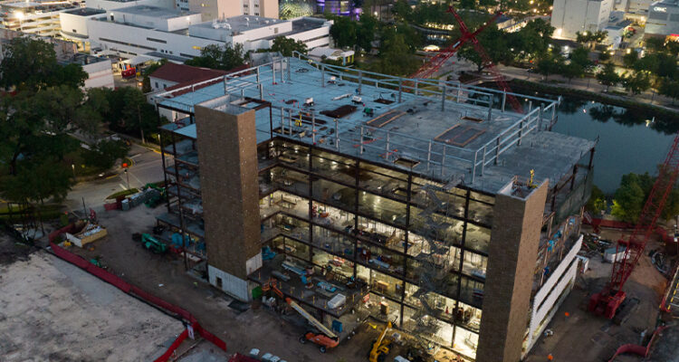 Steel-framed building under construction, vehicles nearby, city at dusk.