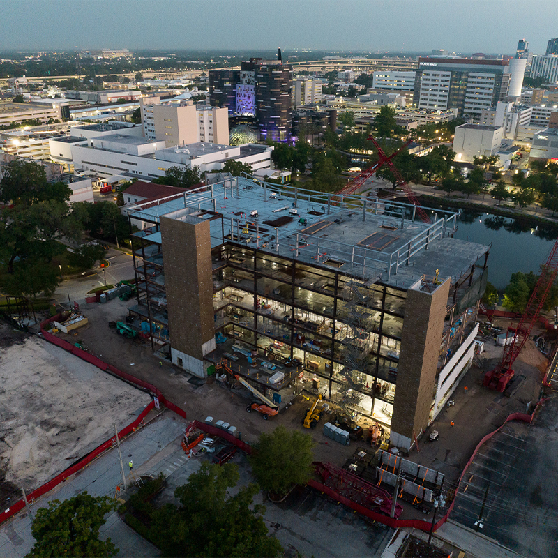 Steel-framed building under construction, vehicles nearby, city at dusk.