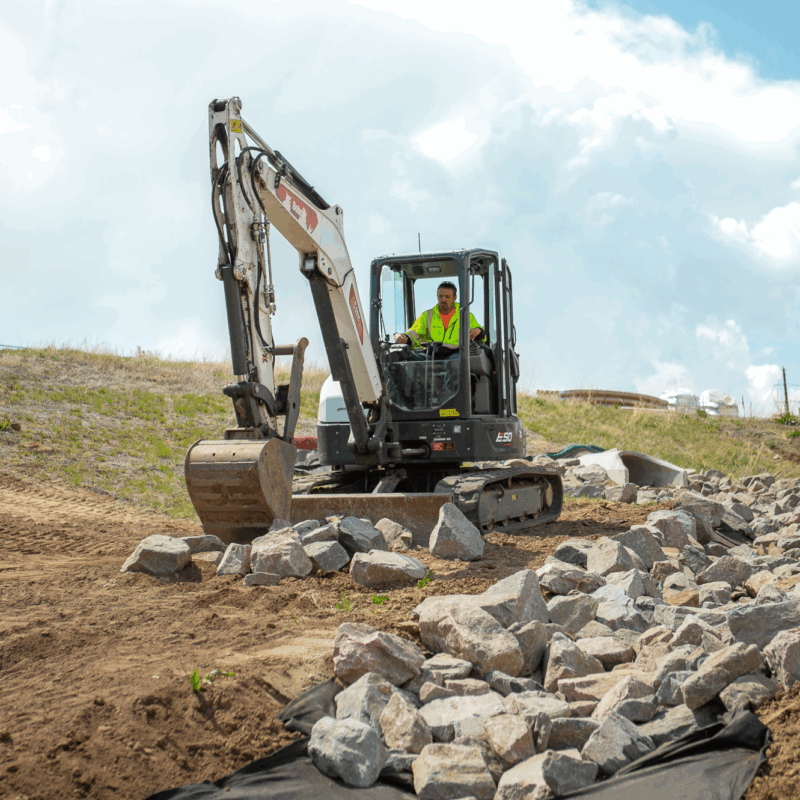 Worker in neon vest uses excavator to move rocks outdoors.