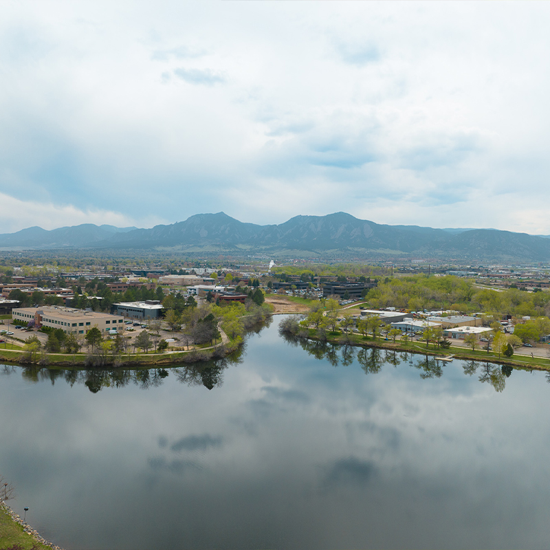 Lake reflects trees, buildings, mountains and a cloudy sky.