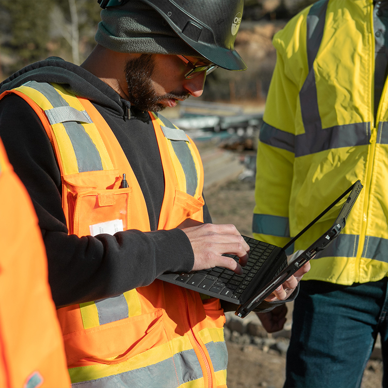Two workers in safety gear use a laptop at construction site.