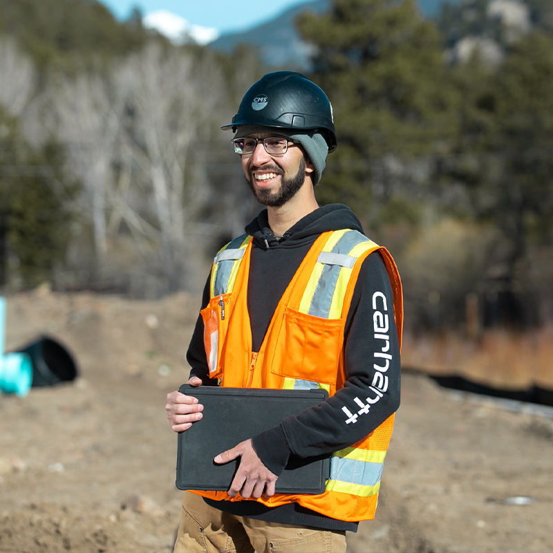 Person in hard hat and vest holds closed laptop at construction site.