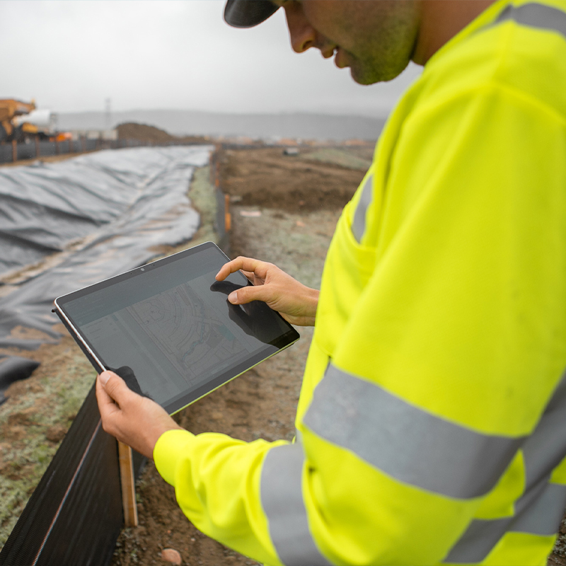 Person in hi-vis jacket uses tablet at construction site.
