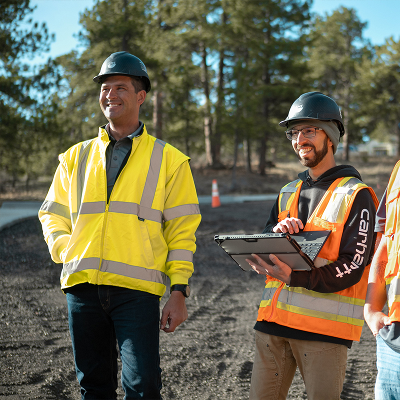 Two construction workers in safety gear stand on a gravel road.