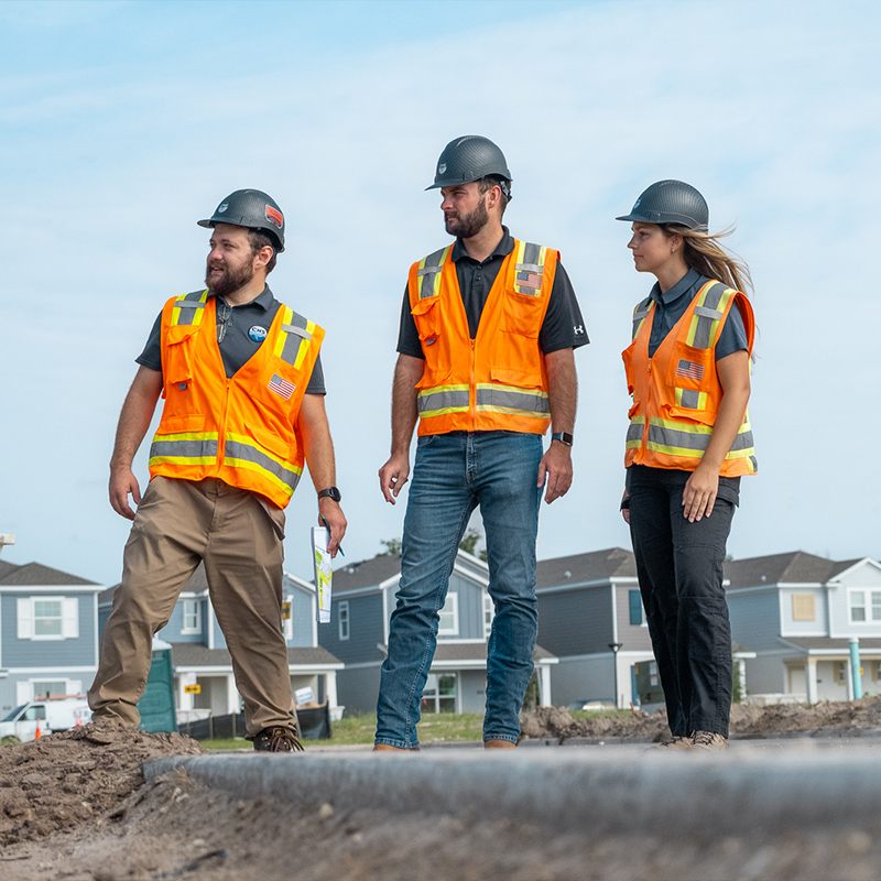 Three construction workers in orange vests and hard hats onsite.