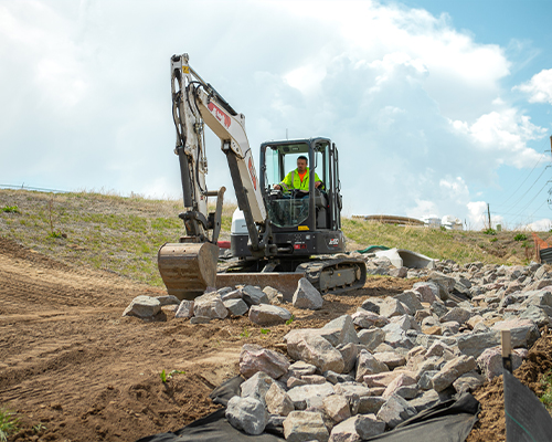 Worker uses small excavator to move rocks on grassy slope.