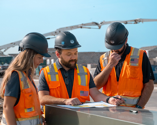 Three construction workers in orange vests review documents outside.