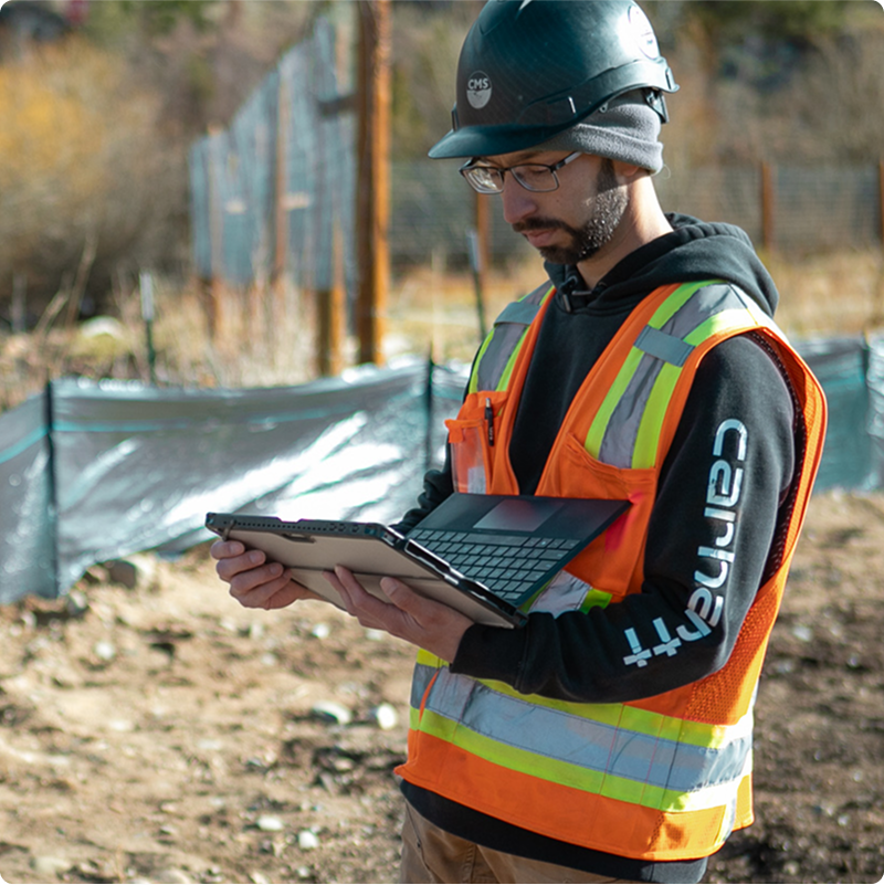 Worker in safety gear uses laptop at construction site.