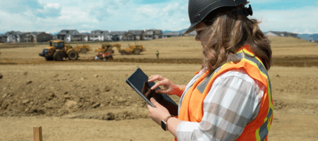 Person in hard hat and vest uses tablet at construction site.