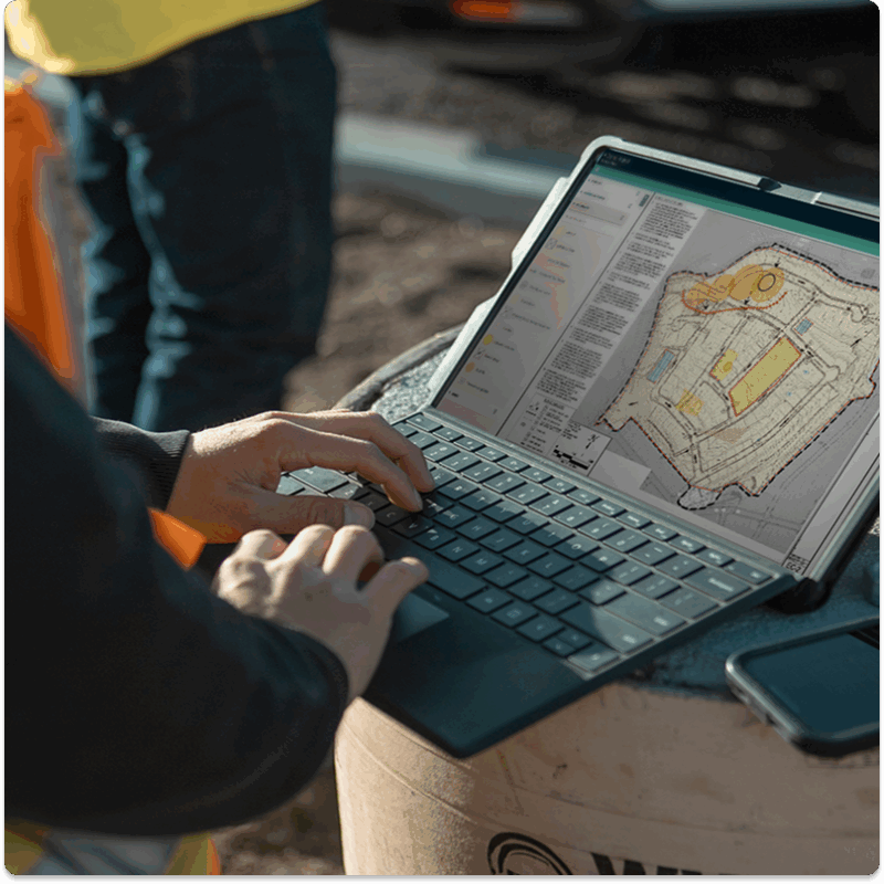 Person on laptop editing site plan outdoors, construction behind.