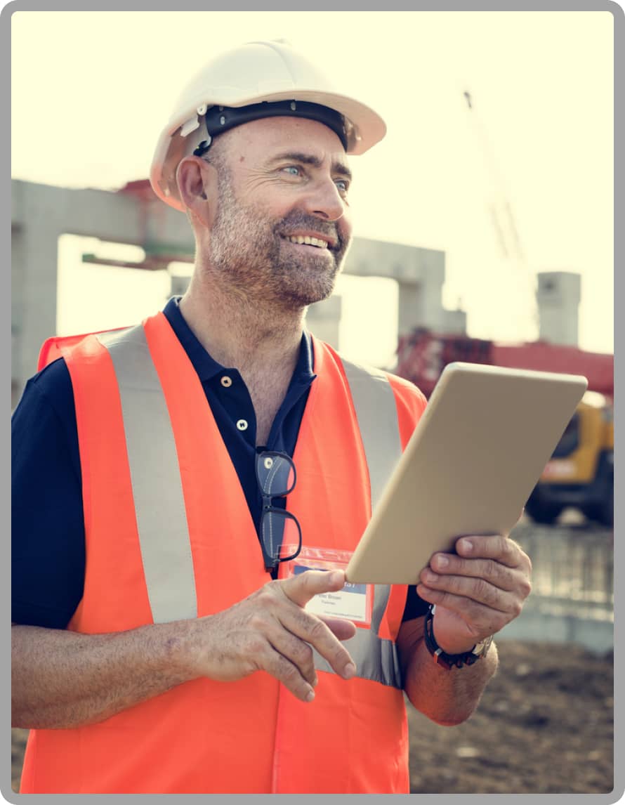 A smiling construction worker in a hard hat and orange vest holds a tablet at a construction site. The scene conveys a sense of productivity and positivity.